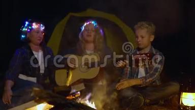 少女一起用吉他唱歌曲，妈妈和哥哥在闪闪发光的篝火背景下。 女孩在演奏音乐